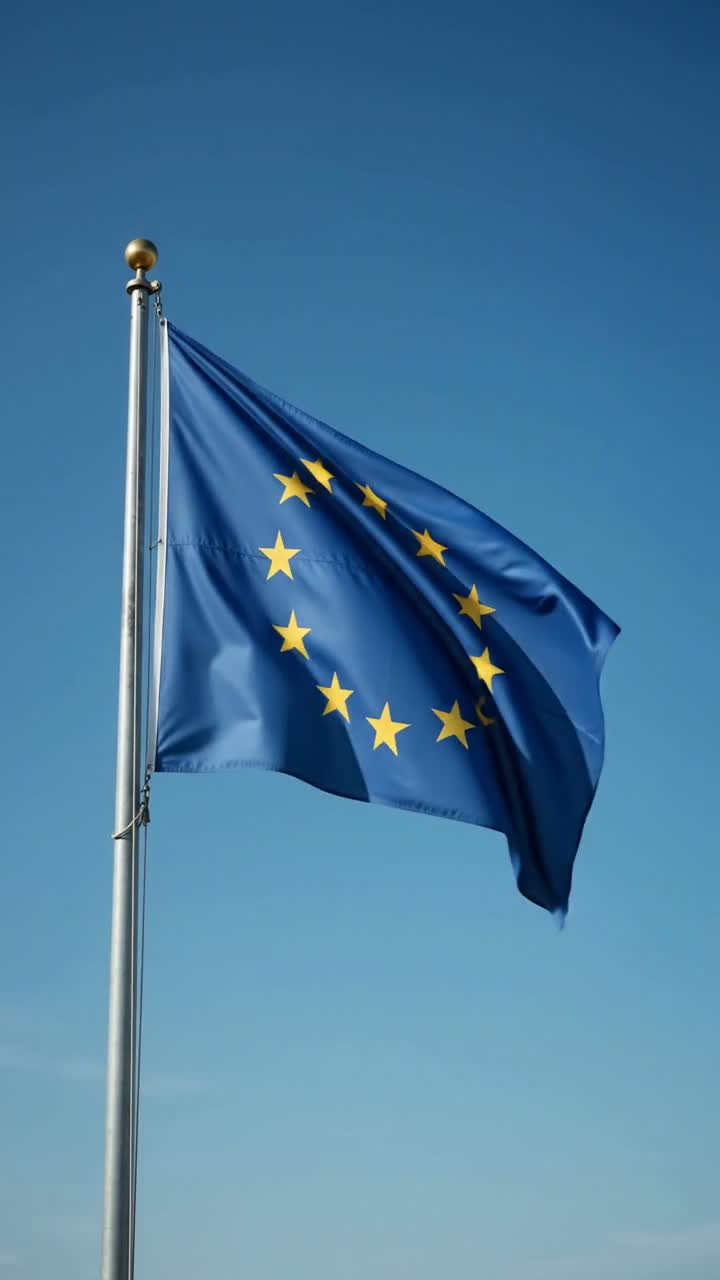 A wide, low-angle static shot frames a European Union flag mounted on a tall metal pole, its twelve gold stars and deep blue field rippling in slow, rhythmic waves against an empty, brilliant azure sky. Soft midday sunlight rakes across the fabric, catching subtle folds and creases as gentle gusts push through, the flag billowing outward then relaxing in a steady, hypnotic cycle. The lens holds perfectly still, shallow depth of field keeping the flag crisp while the cloudless sky remains a clean gradient backdrop. Minimalist, serene, cinematic mood with a calm blue-and-gold palette. Audio: soft continuous wind whoosh and the faint rhythmic flutter and snap of fabric.